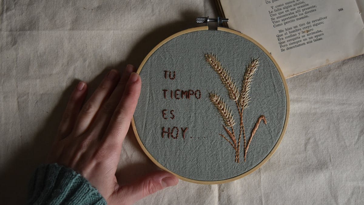 A hand touches an embroidery hoop with Spanish text and flowers beside an open book.